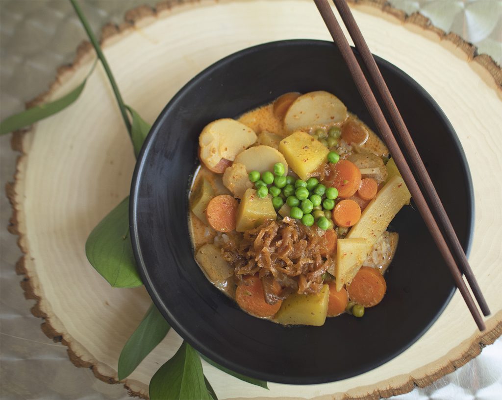 A black bowl of vegan massaman curry with large chunks of potatoes, carrots, and bamboo shoots. There are a pair of wooden chop sticks in the bowl and the bowl is resting on a wooden circle