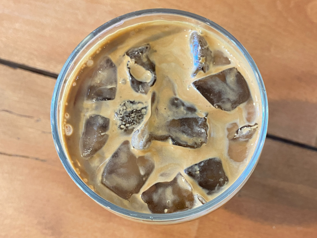 A fresh glass of iced coffee and oat milk sits on a wooden table.